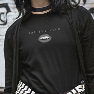 Puede incluir: Una camiseta negra de manga larga con un gr&aacute;fico blanco de una boca de vampiro con colmillos y el texto "eat the rich".