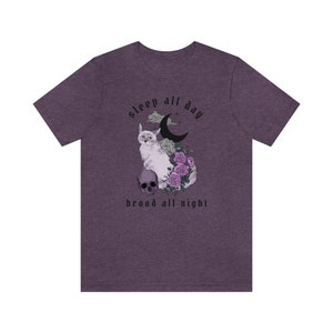 K&ouml;nnte beinhalten: Lilafarbenes T-Shirt aus Heather-Material mit einer wei&szlig;en Katze, einem zunehmenden Mond und Blumen. Der Text "sleep all day brood all night" ist auf das T-Shirt gedruckt.