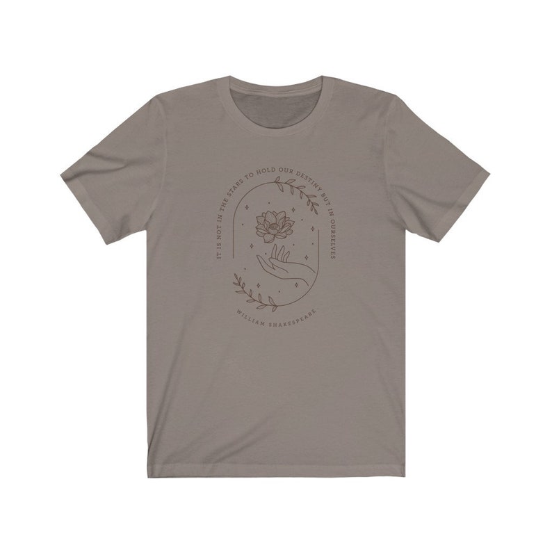 May include: A gray t-shirt with a brown outline of a hand holding a flower with stars around it. The text reads "It is not in the stars to hold our destiny but in ourselves" and "William Shakespeare".