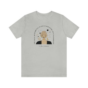 Może przedstawiać: Jasnoszary t-shirt z czarno-złotą ilustracją kobiety z koroną celestialną. Tekst "The fault, dear Brutus, is not in our stars, but in ourselves" jest nadrukowany czarnym kolorem z cytatem Williama Szekspira.