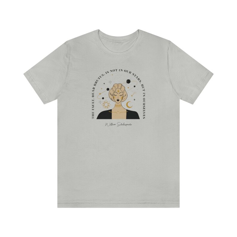 Może przedstawiać: Jasnoszary t-shirt z czarno-złotą ilustracją kobiety z koroną celestialną. Tekst "The fault, dear Brutus, is not in our stars, but in ourselves" jest nadrukowany czarnym kolorem z cytatem Williama Szekspira.