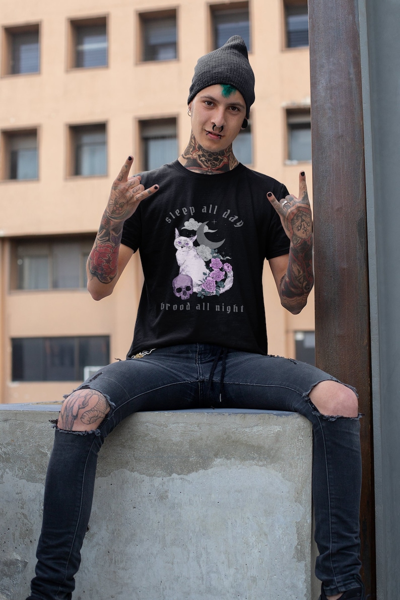 K&ouml;nnte beinhalten: Ein schwarzes T-Shirt mit einem Motiv einer Katze, Mond und Blumen. Der Text auf dem Shirt lautet "sleep all day brood all night".