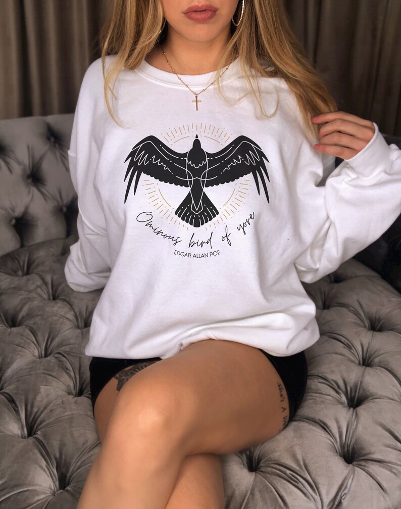 K&ouml;nnte beinhalten: Wei&szlig;er Pullover mit einer schwarzen Silhouette eines Vogels mit dem Text "Ominous bird of yore" und "Edgar Allan Poe" auf der Vorderseite.