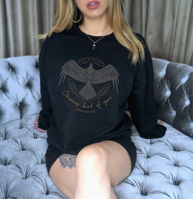 K&ouml;nnte beinhalten: Schwarzer Pullover mit einem goldenen Grafikdruck eines Vogels in einem Kreis mit dem Text "ominous bird of yore" und "Edgar Allan Poe" darunter.