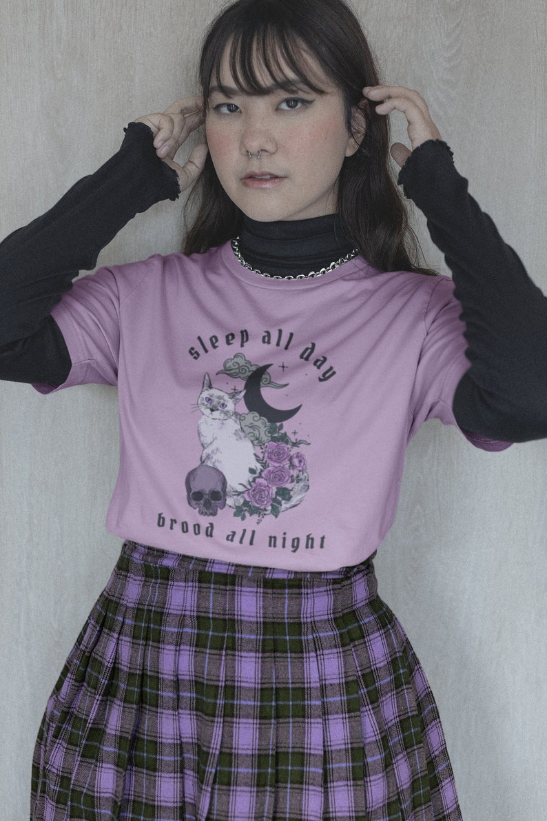 K&ouml;nnte beinhalten: Ein lila T-Shirt mit einem Aufdruck einer Katze, eines Mondes und Blumen. Der Text auf dem Shirt lautet "sleep all day brood all night".
