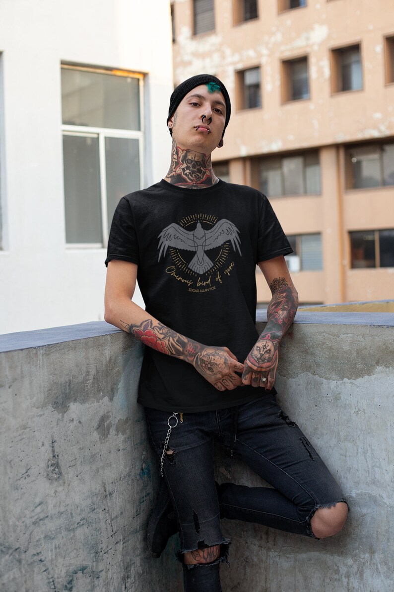 Puede incluir: Una camiseta negra con un gr&aacute;fico de un p&aacute;jaro en vuelo con el texto "Ominous bird of you" y "Edgar Allan Poe" debajo. La persona que lleva la camiseta tiene tatuajes en los brazos y el cuello.