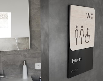 Op de afbeelding: Een zwart en houten bord met de tekst "WC" en "Toalet" in witte letters. Het bord bevat ook een pictogram van een man, een vrouw en een rolstoelgebruiker.