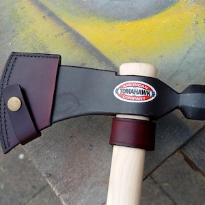 May include: A black metal tomahawk with a light wood handle, featuring a dark burgundy leather blade sheath with a brass snap button and a matching leather collar. The head displays an oval sticker reading American Tomahawk Company.
