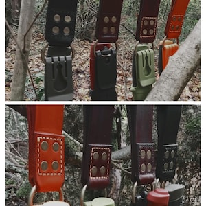 May include: A collection of leather knife sheaths in various colors, including black, brown, and orange, are displayed. Each sheath features a metal ring and is attached to a knife. The sheaths are secured with rivets and are hanging from a wooden branch.