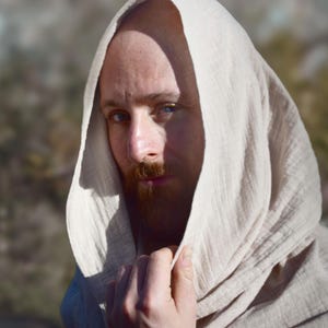 Könnte beinhalten: Eine Person trägt ein hellbeige Kapuzengewand mit rotem Bart und blauen Augen. Der Stoff der Kapuze ist strukturiert und um Kopf und Schultern drapiert. Die Person schaut direkt in die Kamera.
