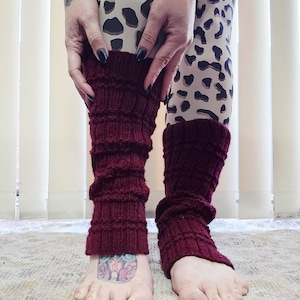May include: Burgundy-colored knitted leg warmers. The leg warmers are made of a thick, textured knit and are worn over the lower legs. The person in the image is wearing leopard print leggings. The leg warmers are pulled up to the knees.