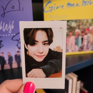 May include: A Polaroid photo of a young man with dark hair. He is wearing a black sweater and looking at the camera. The photo is held in a hand with pink nail polish.