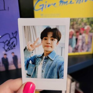 May include: A white Polaroid photo of a young man with dark hair wearing a blue denim jacket and a white shirt. He is smiling and making a peace sign with his fingers. The photo is on a bookshelf with other books and a yellow album with the text "Give Me More" on the cover.