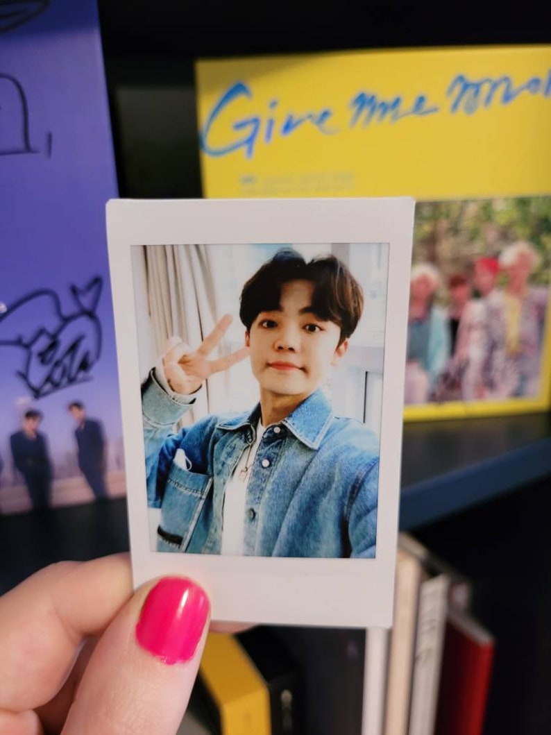 May include: A white Polaroid photo of a young man with dark hair wearing a blue denim jacket and a white shirt. He is smiling and making a peace sign with his fingers. The photo is on a bookshelf with other books and a yellow album with the text "Give Me More" on the cover.