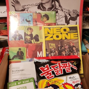 May include: A box filled with a variety of K-pop merchandise, including albums, photo cards, and snacks. The albums feature the groups NCT, Ateez, and Stray Kids. The photo cards feature members of the groups NCT and Stray Kids. The snacks are a bag of spicy Korean ramen noodles.