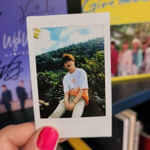 May include: A Polaroid photo of a person wearing a white t-shirt with an orange design and white pants sitting on a rock in front of a green forest. The photo is held in a hand with pink nail polish.