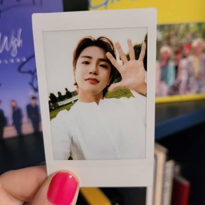 May include: A Polaroid photo of a young man with brown hair wearing a white shirt. He is smiling and holding his hand up to the camera. The photo is held in a hand with pink nail polish.