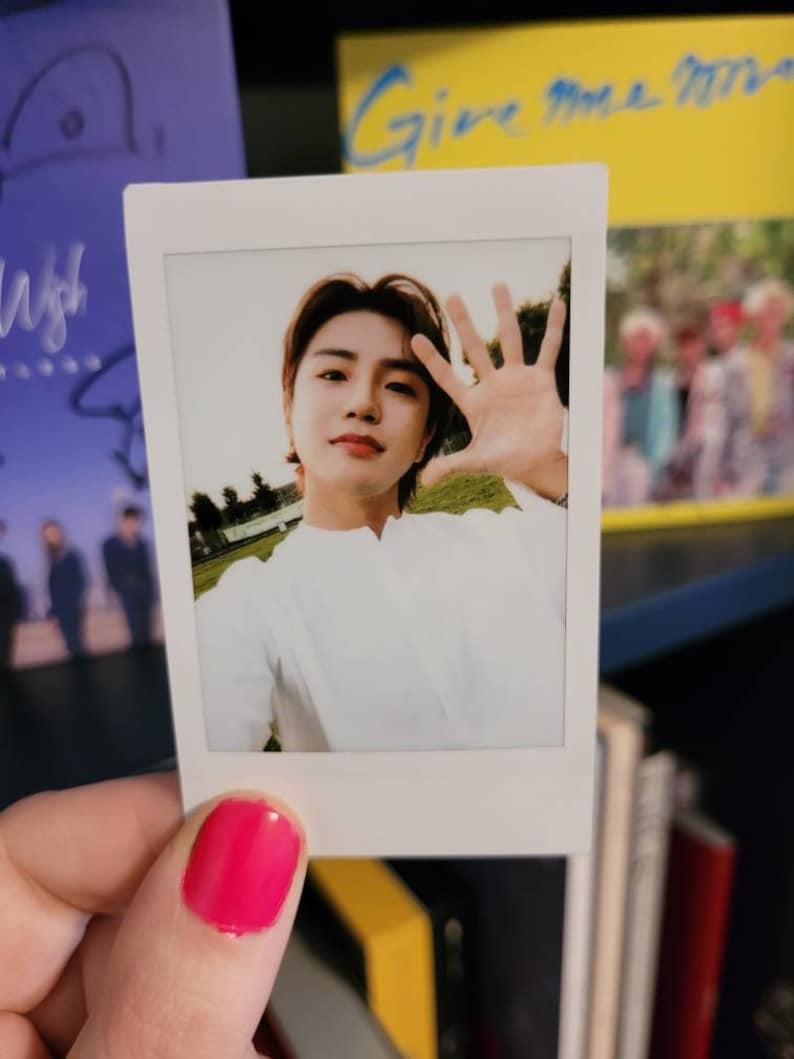 May include: A Polaroid photo of a young man with brown hair wearing a white shirt. He is smiling and holding his hand up to the camera. The photo is held in a hand with pink nail polish.