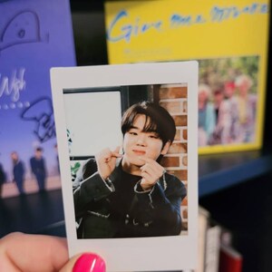 May include: A Polaroid photo of a young man with dark hair, wearing a black jacket, making a heart shape with his hands. The photo is held in a hand with pink nail polish.