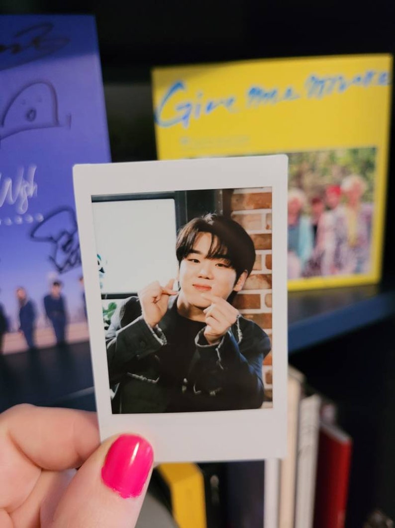 May include: A Polaroid photo of a young man with dark hair, wearing a black jacket, making a heart shape with his hands. The photo is held in a hand with pink nail polish.