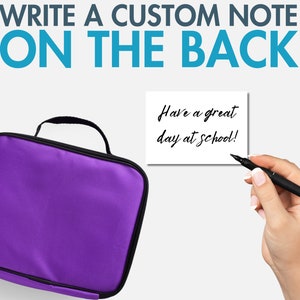 Peut inclure: Un sac &agrave; d&eacute;jeuner violet avec une poign&eacute;e noire et une fermeture &eacute;clair. Le sac a une note blanche vierge attach&eacute;e &agrave; lui avec le texte "Have a great day at school!" &eacute;crit dessus.