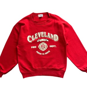 Könnte beinhalten: Ein rotes Sweatshirt mit Rundhalsausschnitt und dem Wort "CLEVELAND" in cremefarbener Bogenform. Darunter steht "FIRE DEPT." und ein Feuerwehrabzeichen. Das Sweatshirt trägt auch den Text "PROUD TO SERVE".