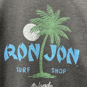 May include: Dark grey crew neck sweatshirt featuring a palm tree graphic and the words "RON JON SURF SHOP Orlando" in blue and white. The sweatshirt is on a wooden hanger.