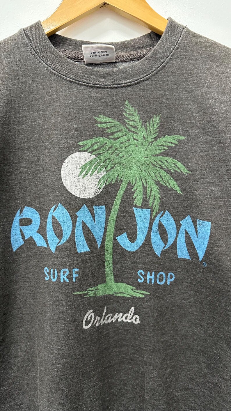 May include: Dark grey crew neck sweatshirt featuring a palm tree graphic and the words "RON JON SURF SHOP Orlando" in blue and white. The sweatshirt is on a wooden hanger.