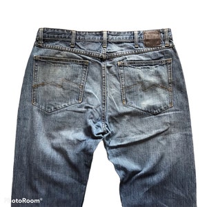 Peut inclure: Vue arri&egrave;re d'un jean en denim bleu avec deux poches arri&egrave;re et un patch en cuir marron. Le jean a une conception classique &agrave; cinq poches et une fermeture &agrave; bouton et &agrave; glissi&egrave;re. Le denim est d&eacute;lav&eacute; et us&eacute;.