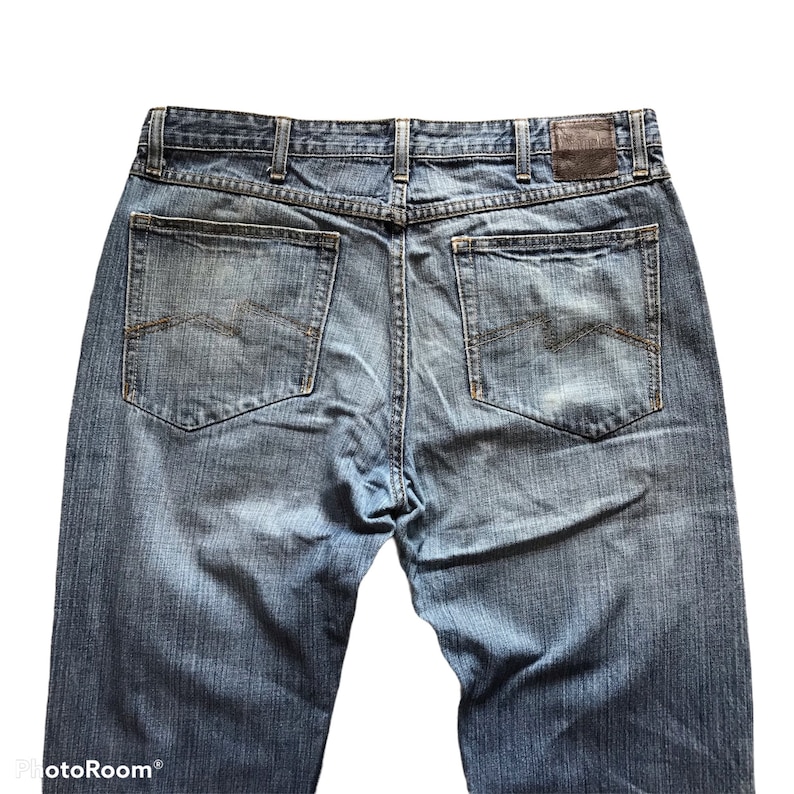 Peut inclure: Vue arri&egrave;re d'un jean en denim bleu avec deux poches arri&egrave;re et un patch en cuir marron. Le jean a une conception classique &agrave; cinq poches et une fermeture &agrave; bouton et &agrave; glissi&egrave;re. Le denim est d&eacute;lav&eacute; et us&eacute;.