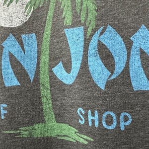 May include: Dark grey t-shirt featuring a green palm tree and a white moon graphic. The text "NJOR" in blue, and "SHOP" in blue, with "Orlando" in white, is also printed on the shirt.