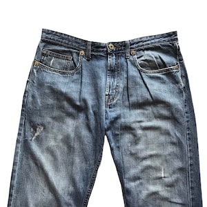 Peut inclure: Gros plan sur un jean en denim bleu avec une fermeture boutonn&eacute;e et des poches. Le jean a un aspect us&eacute; et vieilli avec des zones d&eacute;lav&eacute;es et une petite d&eacute;chirure sur la jambe gauche. Le denim est de couleur moyenne.