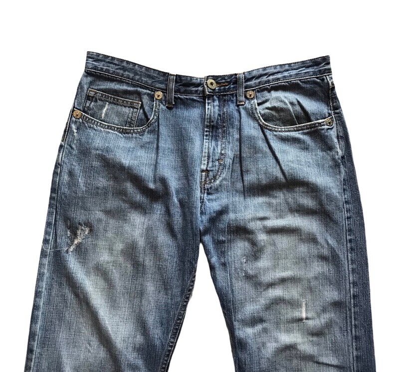 Peut inclure: Gros plan sur un jean en denim bleu avec une fermeture boutonn&eacute;e et des poches. Le jean a un aspect us&eacute; et vieilli avec des zones d&eacute;lav&eacute;es et une petite d&eacute;chirure sur la jambe gauche. Le denim est de couleur moyenne.