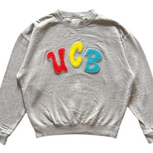 May include: A gray sweatshirt with the letters "UCB" in red, yellow, and blue. The letters are in a playful, bubble-style font and are stitched onto the front of the sweatshirt. The sweatshirt has a crew neck and long sleeves.