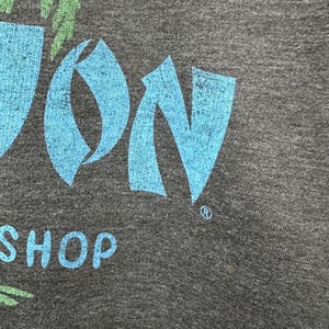 May include: Close-up of a dark grey t-shirt with a teal and green graphic. The graphic includes the word "JON" in teal, a palm tree design, and the word "SHOP" in white. The shirt has a textured appearance.