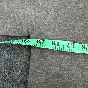 May include: Close-up of a grey t-shirt with a green measuring tape. The tape shows measurements from 15 to 51 cm. The fabric has a textured appearance.