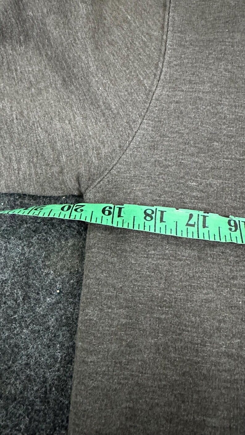 May include: Close-up of a grey t-shirt with a green measuring tape. The tape shows measurements from 15 to 51 cm. The fabric has a textured appearance.