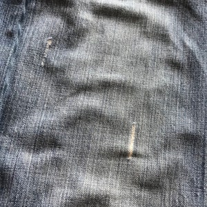 Peut inclure: Gros plan sur un tissu denim avec une texture visible et des nuances subtiles. Le tissu du jean bleu pr&eacute;sente des signes d'usure, dont une petite d&eacute;chirure r&eacute;par&eacute;e. L'aspect g&eacute;n&eacute;ral est d&eacute;lav&eacute; et textur&eacute;.
