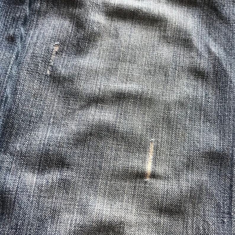 Peut inclure: Gros plan sur un tissu denim avec une texture visible et des nuances subtiles. Le tissu du jean bleu pr&eacute;sente des signes d'usure, dont une petite d&eacute;chirure r&eacute;par&eacute;e. L'aspect g&eacute;n&eacute;ral est d&eacute;lav&eacute; et textur&eacute;.