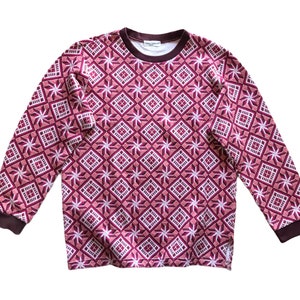 Puede incluir: Una sudadera de manga larga con un estampado geométrico en tonos burdeos, rosa y blanco. El diseño presenta formas de diamantes y estrellas. El cuello y los puños son de color marrón oscuro. La sudadera es de material suave.