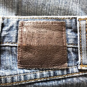 Peut inclure: Gros plan sur une &eacute;tiquette de jean en denim. L'&eacute;tiquette en cuir marron indique "PREMIUM DENIM Wrangler Jeans Co. Since 1947". Le jean est d&eacute;lav&eacute; bleu clair avec des coutures visibles.