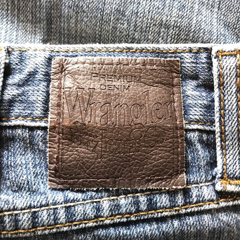 Peut inclure: Gros plan sur une &eacute;tiquette de jean en denim. L'&eacute;tiquette en cuir marron indique "PREMIUM DENIM Wrangler Jeans Co. Since 1947". Le jean est d&eacute;lav&eacute; bleu clair avec des coutures visibles.
