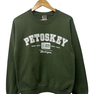 May include: A dark green sweatshirt with the word "PETOSKEY" in white arched lettering. Below the text are the words "GREAT LAKES EST. 1879 GREAT TIMES" and "Michigan" in white. The sweatshirt is hanging on a wooden hanger.