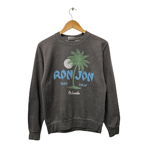 May include: Dark grey sweatshirt with a crew neck and long sleeves. The front displays a graphic of a palm tree, a full moon, and the words "RON JON SURF SHOP Orlando" in blue and green.