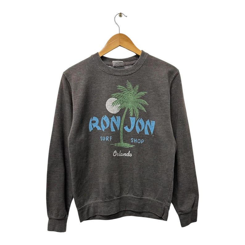 May include: Dark grey sweatshirt with a crew neck and long sleeves. The front displays a graphic of a palm tree, a full moon, and the words "RON JON SURF SHOP Orlando" in blue and green.