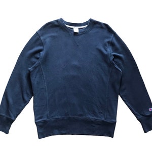 Puede incluir: Una sudadera Champion azul marino con cuello redondo y mangas largas. La sudadera tiene puños acanalados y un pequeño logotipo de Champion en la manga izquierda. La tela parece ser un material suave y cómodo.