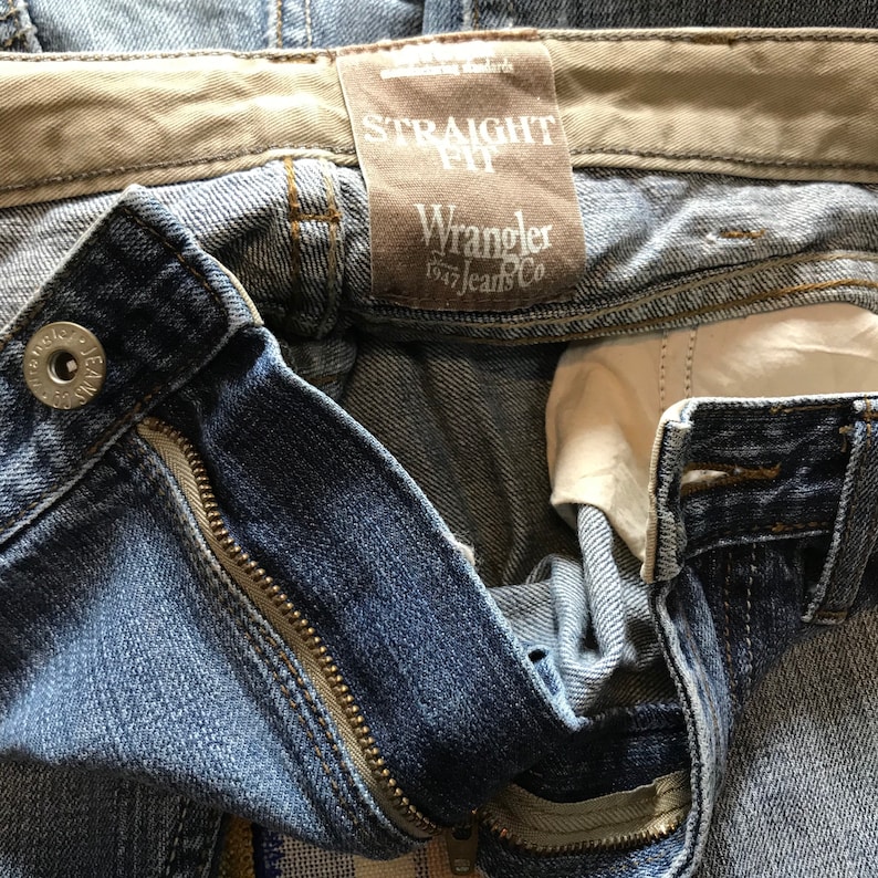 Peut inclure: Gros plan sur un jean en denim bleu avec une fermeture &eacute;clair et un bouton. Le jean porte une &eacute;tiquette marron avec l'inscription "STRAIGHT FIT" et "Wrangler 1947 Jeans Co.". Le jean est l&eacute;g&egrave;rement d&eacute;lav&eacute; et us&eacute;.