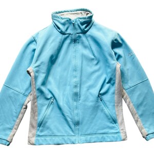 May include: A light blue zip-up jacket with a high collar and two front pockets. The jacket has gray side panels and a gray interior lining. The jacket has a small logo on the upper left sleeve.