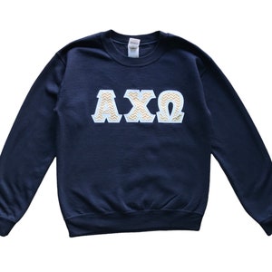 Op de afbeelding: Marineblauw sweatshirt met de Griekse letters ΑΧΩ in een wit en goud chevron patroon. De letters zijn groot en gecentreerd op de borst van de lange mouwen crew neck sweatshirt.
