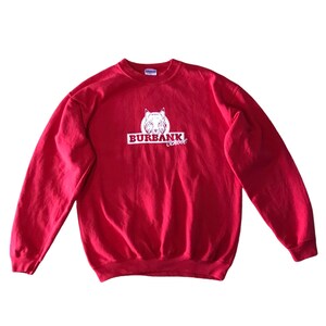 Puede incluir: Sudadera roja de cuello redondo con el texto "BURBANK school" en blanco y un gráfico de lince blanco. La sudadera es de color rojo liso y tiene mangas largas.